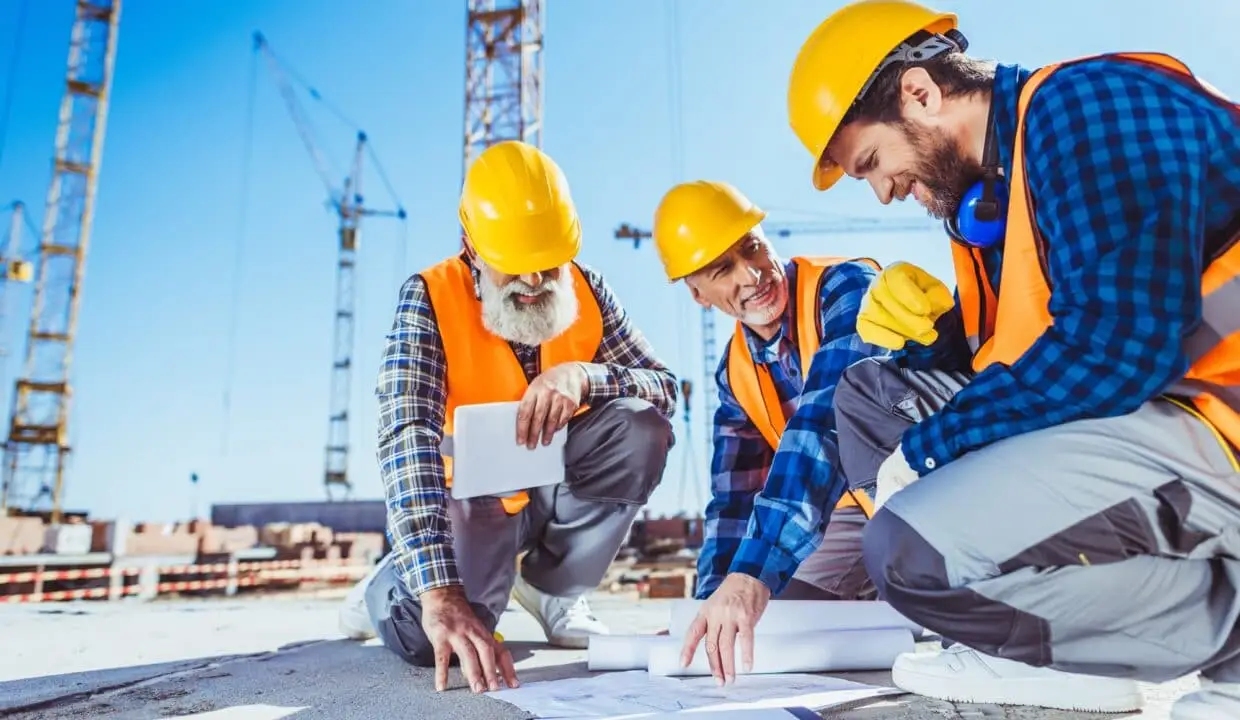 Three construction workers sitting on concrete at construction site, discussing building plans