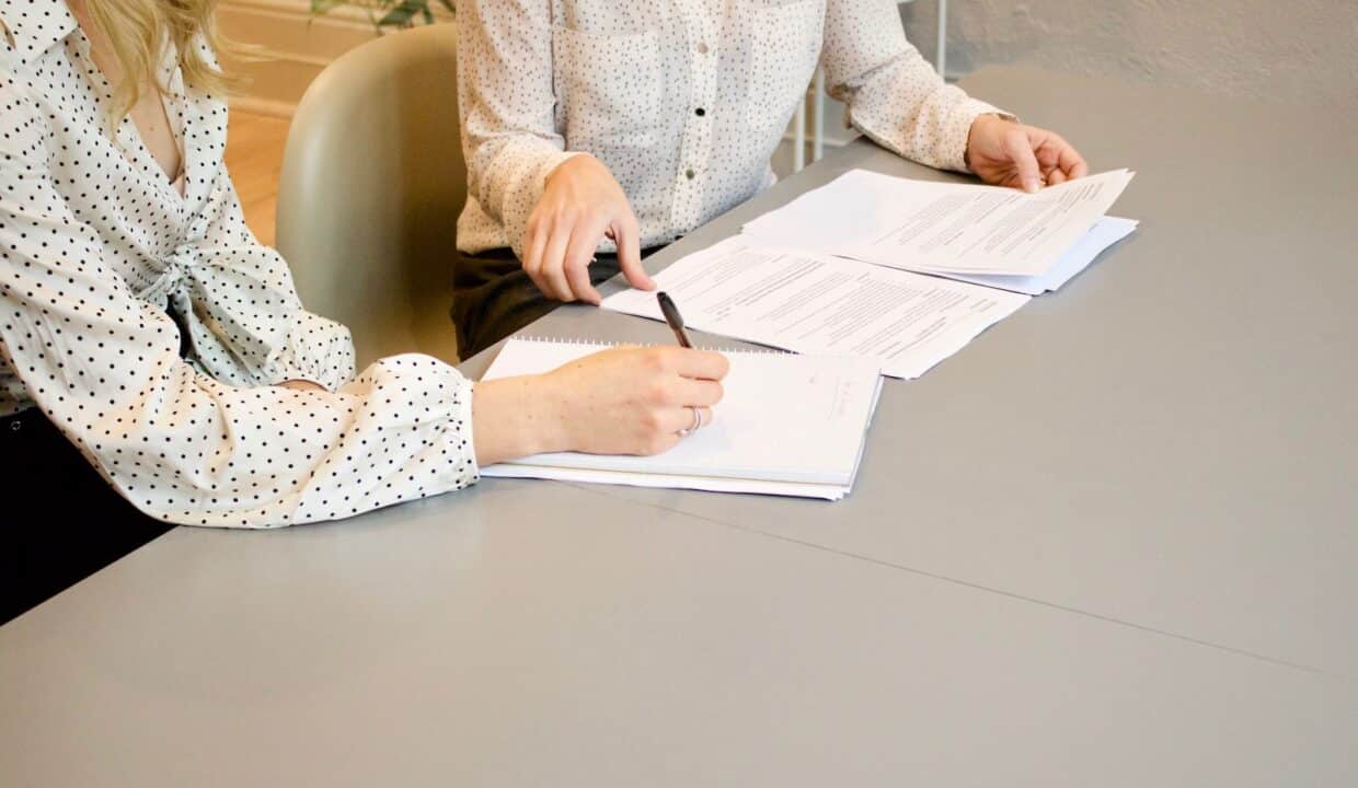 woman signing on white printer paper beside woman about to touch the documents